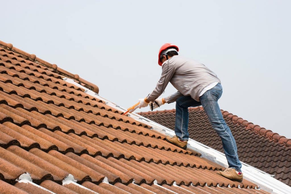 A roofer wearing a safety helmet works on a tiled roof, applying sealant along the roof tiles to repair a leak.